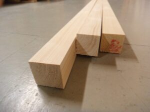 Drei lange Holzleisten liegen auf einem Boden in einer leicht schräge Perspektive; die Maserung des Holzes ist gut sichtbar.
