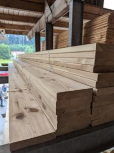 Holzbretter stapeln sich in einem Lager mit Holzdach im Hintergrund.