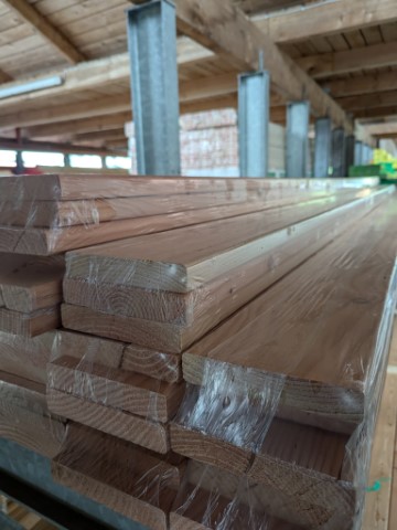 Ein Stapel von Holzplanken, die auf einem Regal in einem Lagerhaus gelagert sind. Der Raum hat eine Holzbalkendecke und Metallstützen.