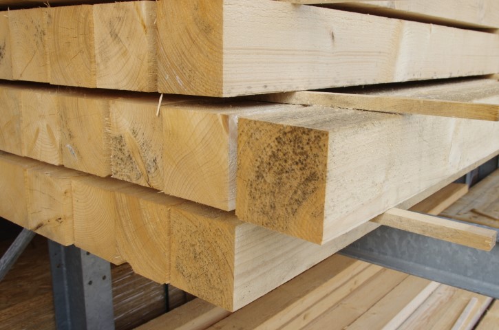Gestapelte Holzbohlen in einem Lager, die eine gleichmäßige Kantenbearbeitung aufweisen und mit Holzleisten voneinander getrennt sind.