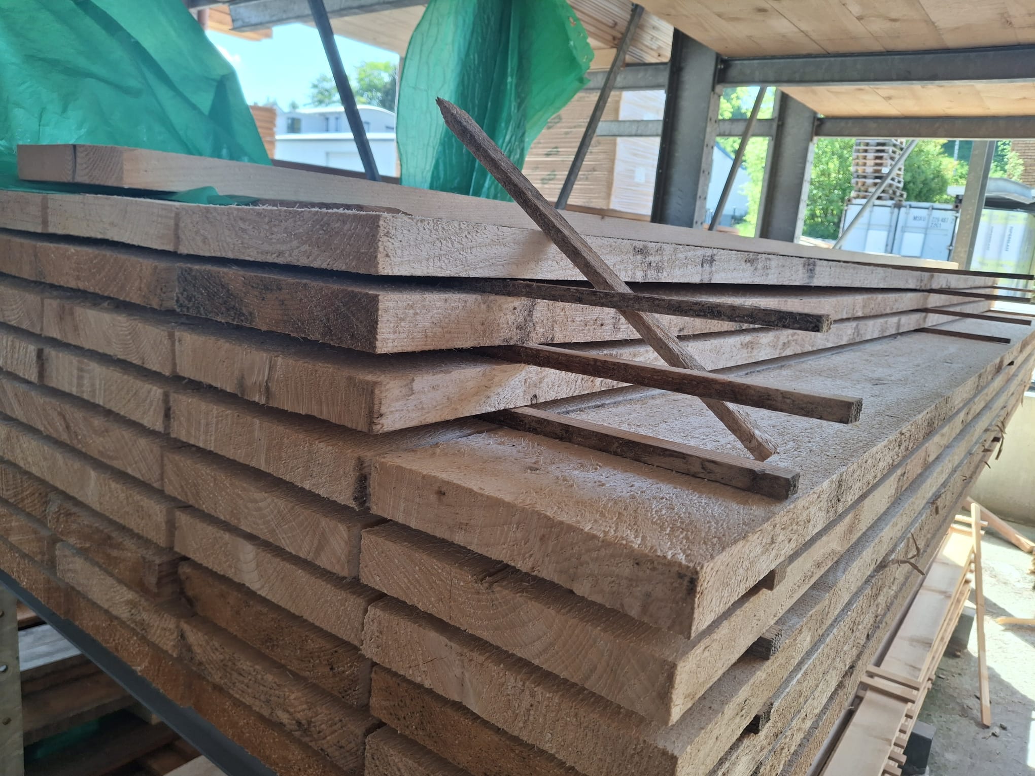 Stapel von Holzbrettern, teils unter grüner Plane auf einer Baustelle gelagert.