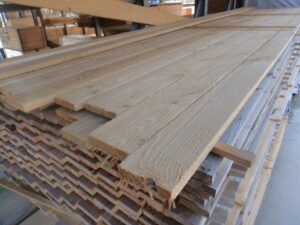 Ein Stapel von Holzbrettern in einem Lager, mit sichtbaren Maserungen und rauen Kanten, die schräg nach oben gestapelt sind.