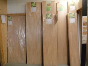 Mehrere Holzplatten unterschiedlicher Größe, aufrecht und in Folie verpackt, stehen nebeneinander an einer Wand, mit Etiketten mit Beschreibungen darauf versehen.