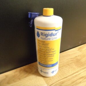 Eine Flasche Rigips Estrichkleber mit gelbem Etikett und Deckel steht auf einem Holztisch vor einem schwarzen Hintergrund.