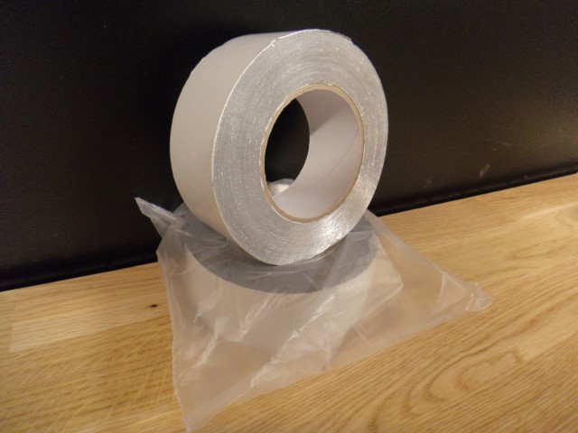 Eine Rolle silbernes Klebeband steht auf einem Holztisch, neben einer transparenten Plastiktüte.