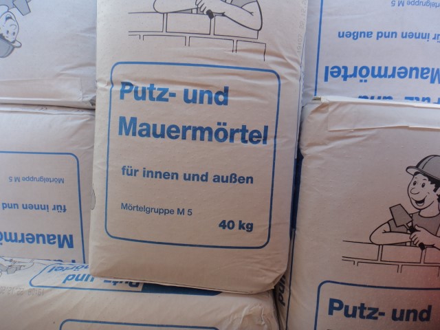 Paket mit der Aufschrift 'Putz- und Mauermörtel für innen und außen, Mörtelgruppe M 5, 40 kg', in einem Stapel von ähnlichen Paketen.