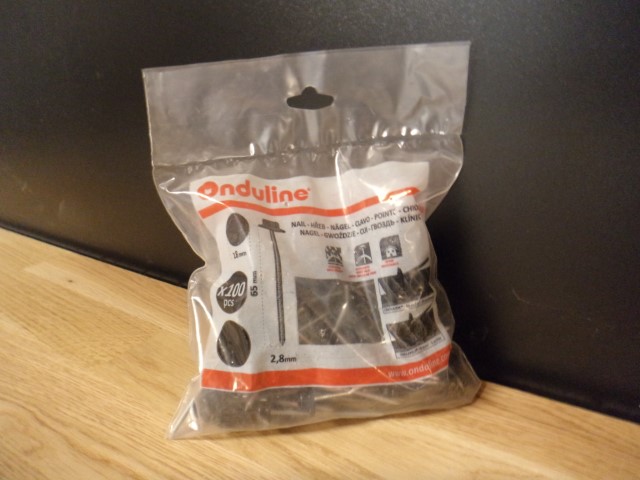 Eine transparente Plastiktüte mit dem Aufdruck 'Onduline', die Nägel enthält, steht auf einem Holztisch vor einem schwarzen Hintergrund. Die Verpackung gibt 200 Stück an und zeigt Abbildungen von Nägeln sowie ihre Dimensionen in Millimetern.