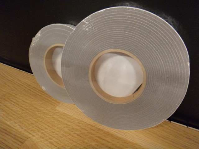 Zwei Rollen silberfarbenes Klebeband nebeneinander auf einem Holzboden liegend, mit einem dunklen Hintergrund.