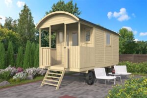 Ein kleiner, holzverkleideter Bauwagen mit gewölbtem Dach steht auf einem gepflasterten Platz. Vor dem Bauwagen befinden sich zwei weiße Gartenstühle und ein kleiner Tisch mit Getränken. Im Hintergrund sind Büsche und Bäume zu sehen, während der Himmel blau und leicht bewölkt ist.