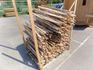 Ein Stapel Holzlatten, sicher gebündelt und auf einer Palette im Freien gelagert, neben einem Holzwagen oder Bauwerk.