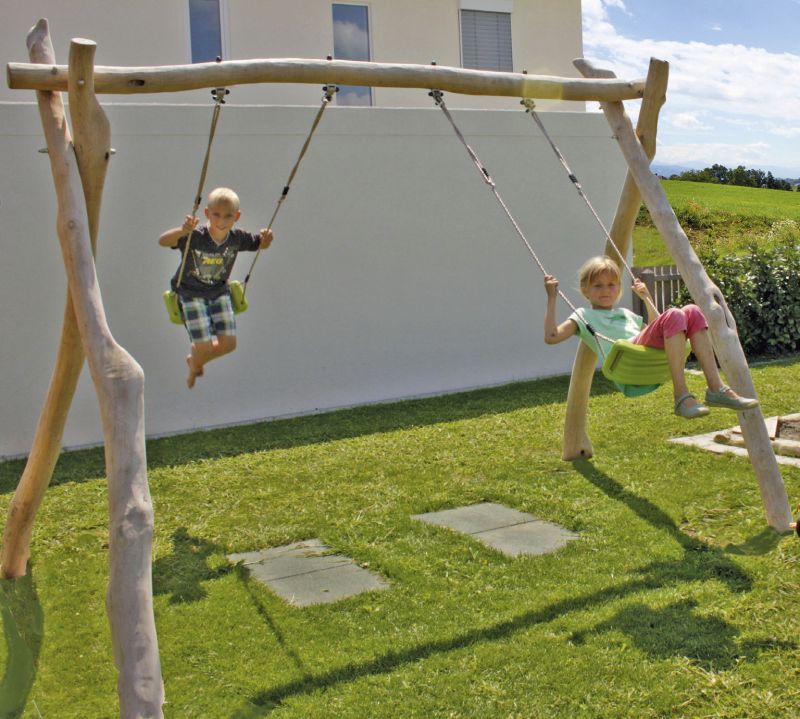 Zwei Kinder schaukeln auf einer Holzschaukel im Garten vor einem weißen Gebäude.