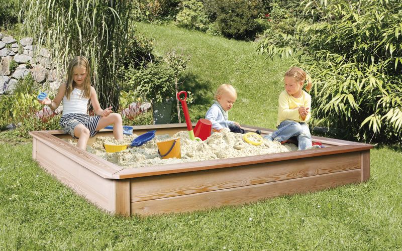 Drei Kinder spielen in einem Sandkasten im Garten, umgeben von grüner Vegetation und Spielzeug.