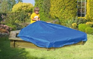 Ein Kind mit einem gelben T-Shirt zieht eine blaue Abdeckung über einen Sandkasten im Garten, umgeben von grünen Hecken und blühenden Pflanzen.
