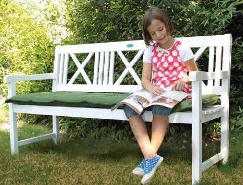Ein Mädchen sitzt auf einer weißen Gartenbank und liest ein Buch. Sie trägt ein weißes T-Shirt mit rosa Punkten und eine kurze Jeanshose. Die Bank steht auf einer Rasenfläche, umgeben von grünen Büschen.