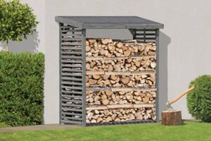 Ein Holzunterstand aus grauem Holz steht an einer weißen Wand und ist voller gestapelter Holzscheite. Daneben liegt ein Holzblock mit einer darin steckenden Axt. Der Boden ist mit grünem Rasen bedeckt, umgeben von Büschen und Bäumen.