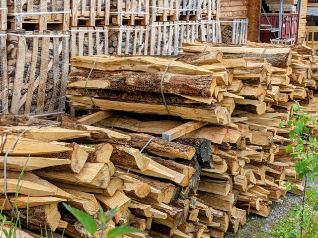Gestapelte Holzscheite, die ordentlich aufeinander geschichtet und mit Gurten gesichert sind. Im Hintergrund sind weitere Holzstapel in einem Lagerbereich zu sehen.