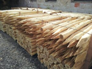 Ein Stapel angespitzter Holzpfähle liegt gestapelt auf einem Boden aus Kieselsteinen neben einer Wand.
