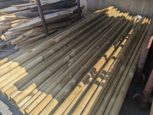 Ein Stapel aus langen, geschälten Holzpfählen, die nebeneinander auf dem Boden liegen, mit Schattenwurf von der Sonne.