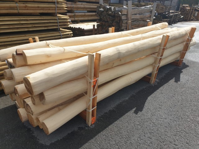 Ein Stapel runder Holzstämme, die mit Bändern auf einem Holzgestell zusammengehalten werden, steht auf einem gepflasterten Boden in einem Außenlager.