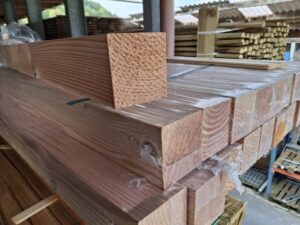 Mehrere Holzbalken sind in einem Lager gestapelt, zum Teil in Plastik eingewickelt. Die Balken haben eine glatte Oberfläche und sind aus hellbraunem Holz. Im Hintergrund sind weitere Holzstapel und ein Teil des Daches zu sehen.