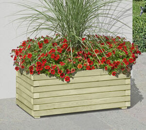 Ein rechteckiger, hölzerner Blumenkasten mit roten Blüten und hohem Ziergras, auf einer gepflasterten Fläche vor einer weißen Wand platziert.