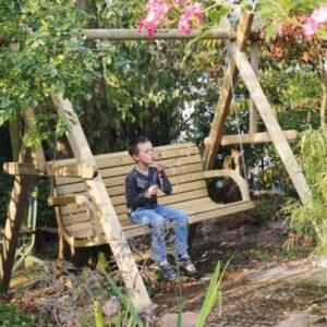 Ein Junge sitzt auf einer Holzschaukel im Garten und bläst Seifenblasen, umgeben von grünen Pflanzen und Blumen.
