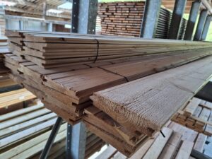 Ein Stapel von Holzbrettern in einer Sägewerksumgebung, gestapelt auf Metallträgern, umgeben von weiteren Holzstapeln im Hintergrund.