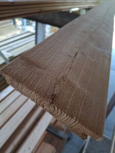 Nahaufnahme eines langen, schmalen Holzbretts mit sichtbarer Maserung, das in einem Lager mit mehreren gelagerten Holzbrettern liegt.