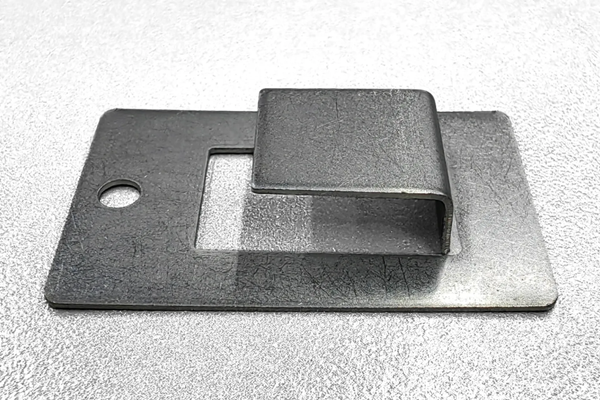 Ein rechteckiges Metallblech mit einem eingeschnittenen, erhobenen Abschnitt in der Mitte, das auf einer glänzenden Oberfläche liegt. Es hat ein rundes Loch auf der linken Seite.