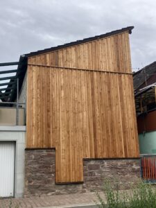 Ein Gebäude mit einer Fassade aus vertikalen Holzlatten und einer unteren Steinmauer, das sich zu einem geneigten Dach erhebt, unter einem bewölkten Himmel.