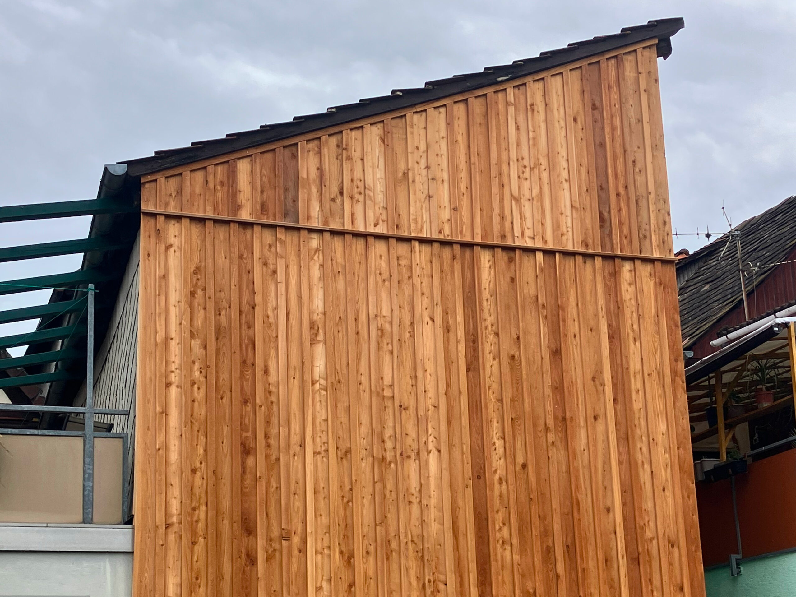 Holzwand eines Hauses mit schrägem Dach, bestehend aus senkrecht angeordneten Holzlatten, im Hintergrund sind weitere Gebäudeteile und ein bewölkter Himmel sichtbar.