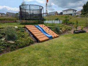 Ein Garten mit einem Trampolin und einem Holzklettergerüst mit Klettergriffen und einer blauen Rutsche, umgeben von Rasen und blühenden Pflanzen.