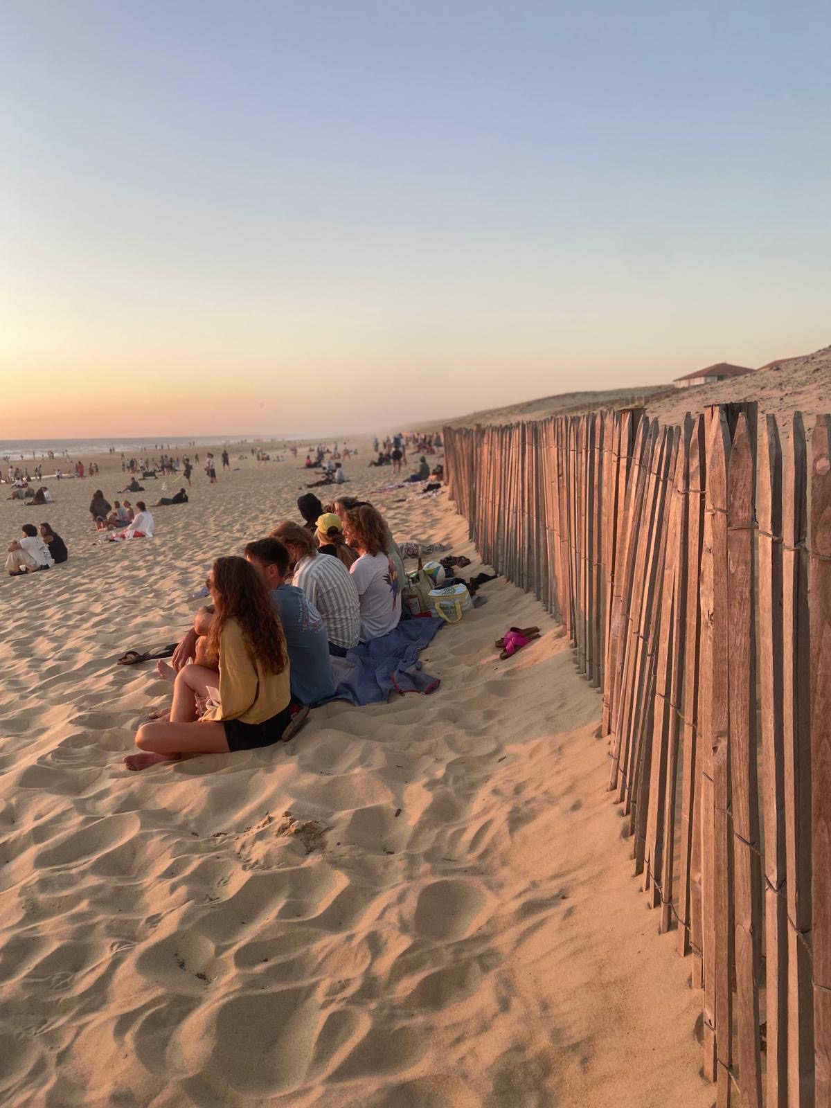 Menschen sitzen am Strand nebeneinander auf einer Decke, mit einem Sandzaun rechts und mehreren anderen Personen im Hintergrund bei Sonnenuntergang.