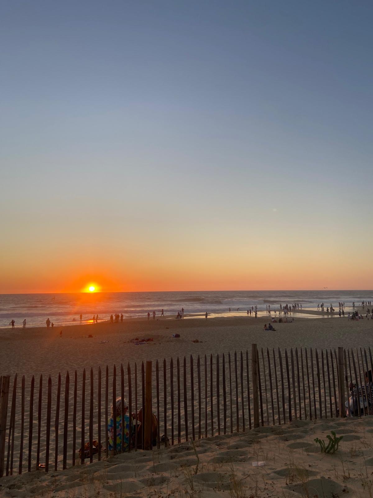 Strandlandschaft bei Sonnenuntergang mit einem Holzzaun im Vordergrund und Menschen, die am Strand spazieren oder sitzen.