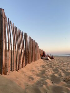 Personen sitzen an einem Sandstrand neben einem Holzzaun bei Sonnenuntergang, im Hintergrund ist das Meer sichtbar.