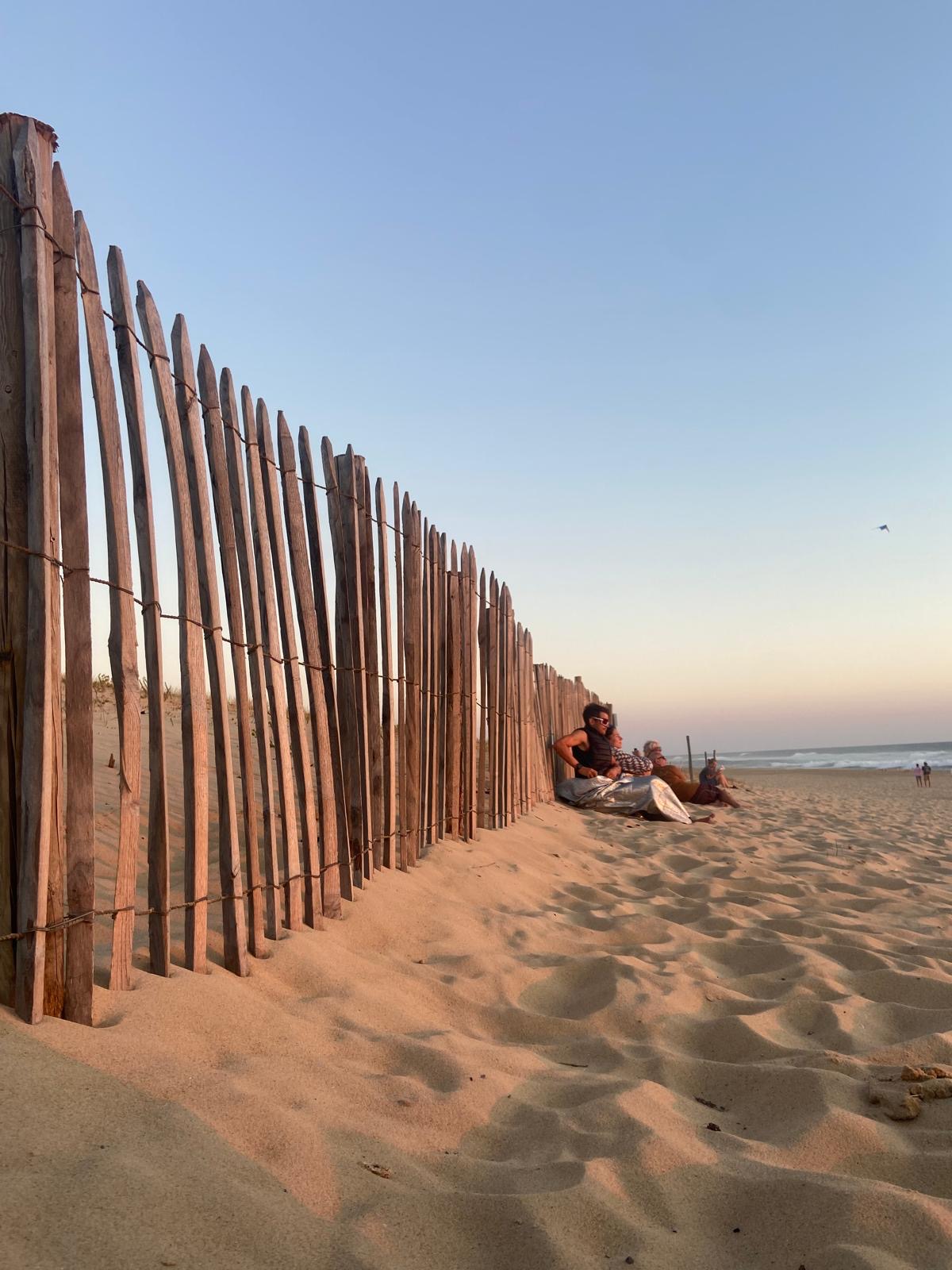 Personen sitzen an einem Sandstrand neben einem Holzzaun bei Sonnenuntergang, im Hintergrund ist das Meer sichtbar.