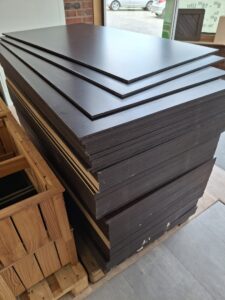 Ein Stapel dunkelbrauner Holzplatten liegt auf einer Palette in einem Innenraum neben einer Holzkiste.