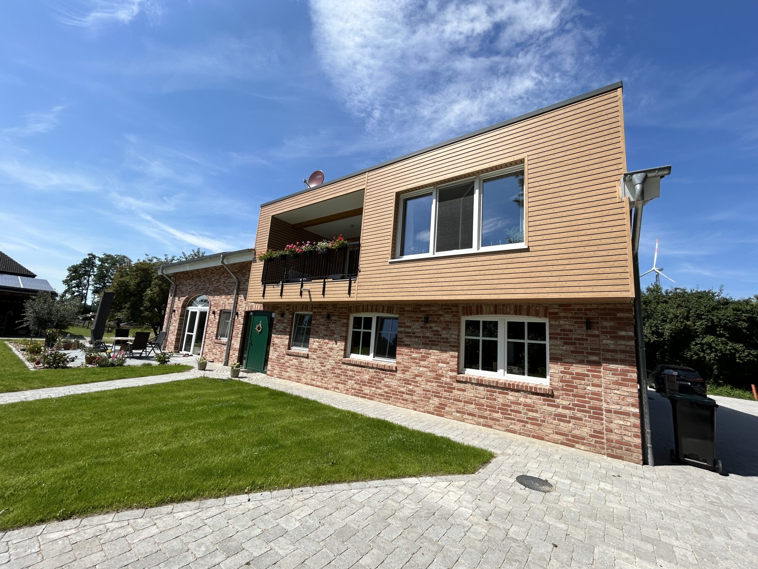 Modernes zweistöckiges Gebäude mit roter Ziegelfassade im Erdgeschoss und Holzverkleidung im Obergeschoss. Vor dem Haus befindet sich ein gepflegter Garten mit Rasenfläche und gepflastertem Weg. Im Hintergrund sind Bäume und ein Windrad zu sehen unter einem blauen Himmel mit leichten Wolken.