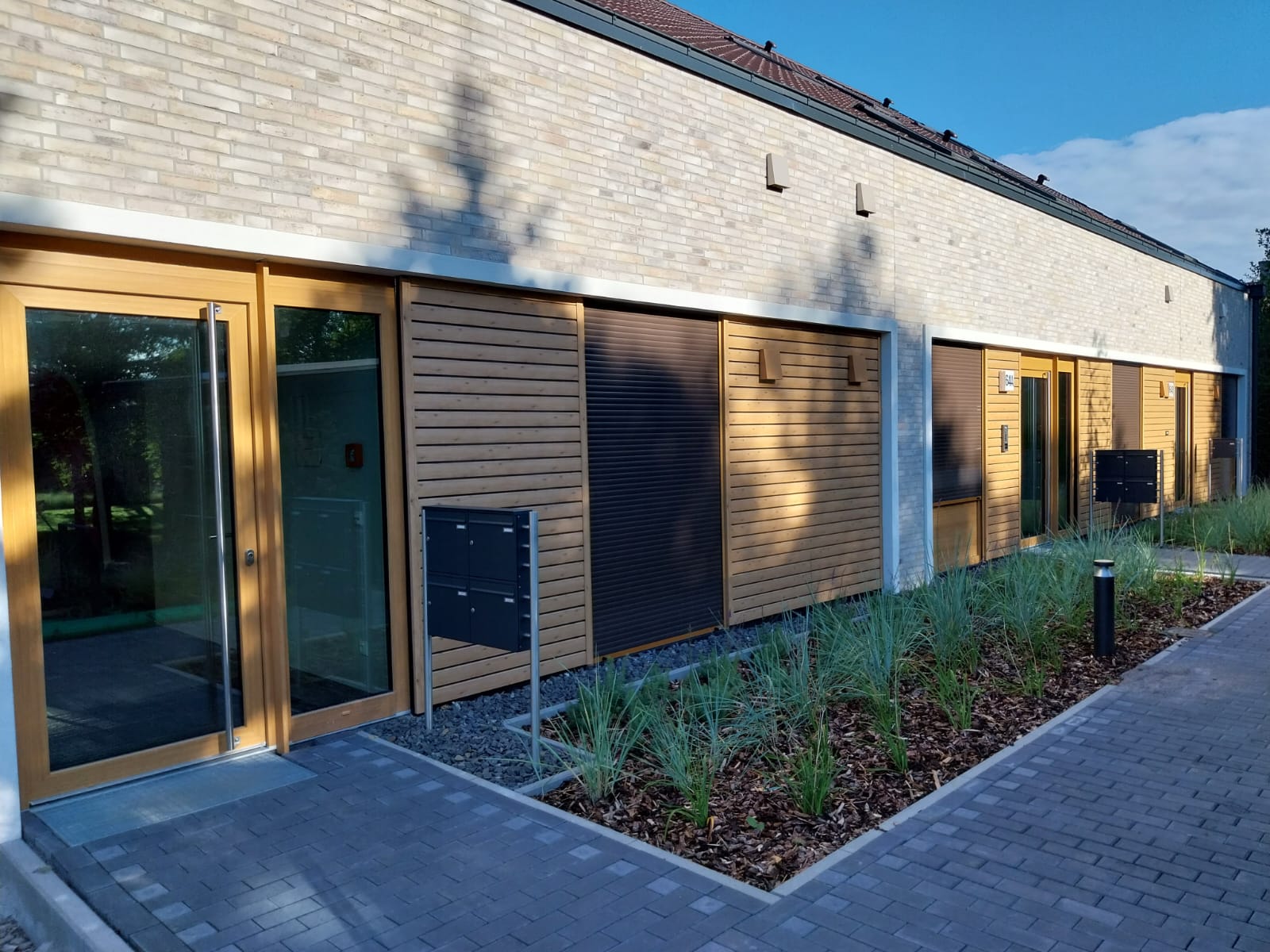 Moderne Gebäudeansicht mit Ziegelaußenwand und Holzverkleidungen, großen Glasfenstern, daneben ein gepflegter Gartenweg mit Kies und Pflanzen.