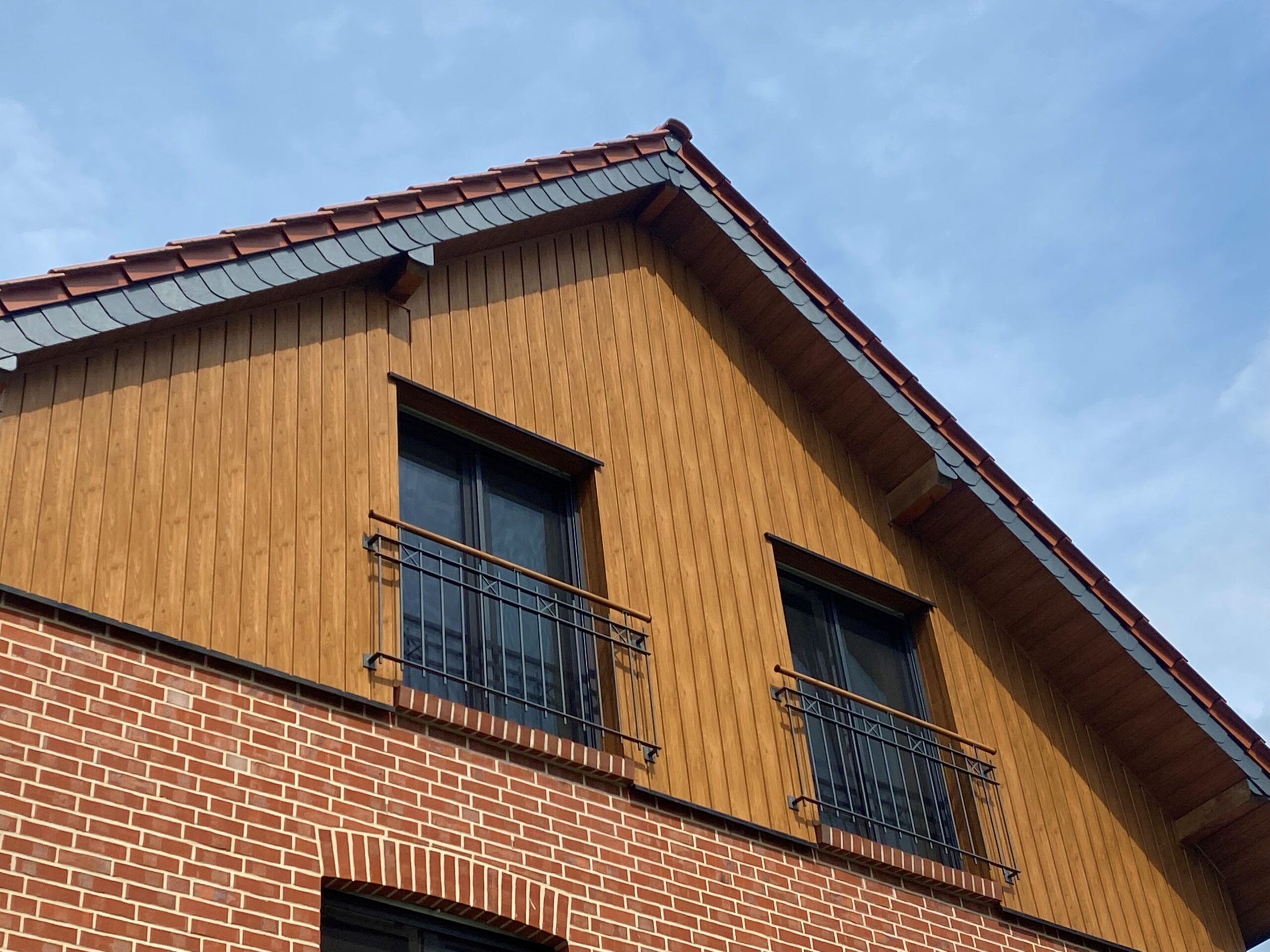 Oberer Teil eines Gebäudes mit einem hölzernen Giebel, zwei Fenstern mit schwarzen Gittern und einer Backsteinfassade darunter, unter einem blauen Himmel.