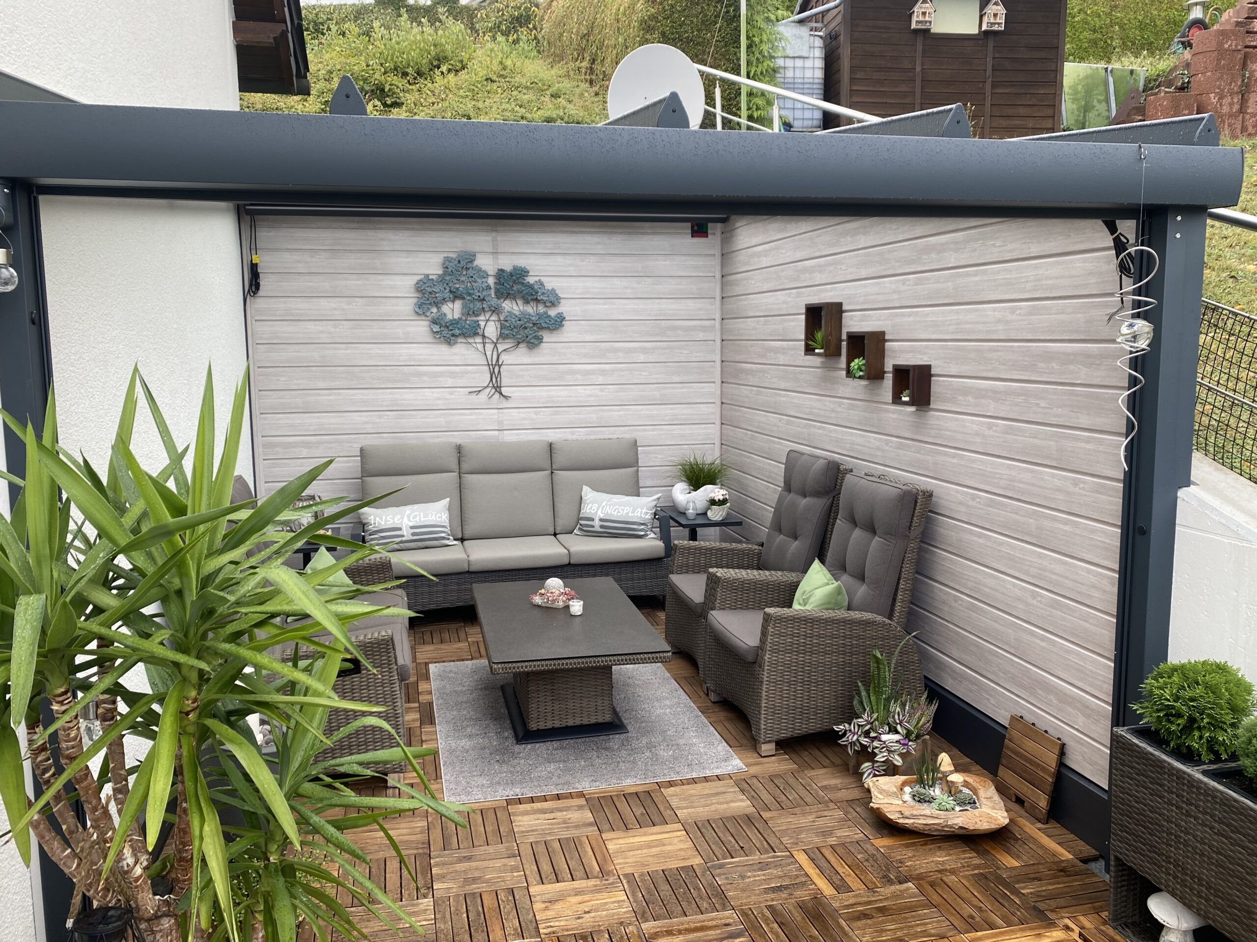 Gemütliche überdachte Terrasse mit grauem Sofa und Sesseln aus Rattan, einem Couchtisch, grüner Pflanze im Vordergrund und moderner Wanddekoration.