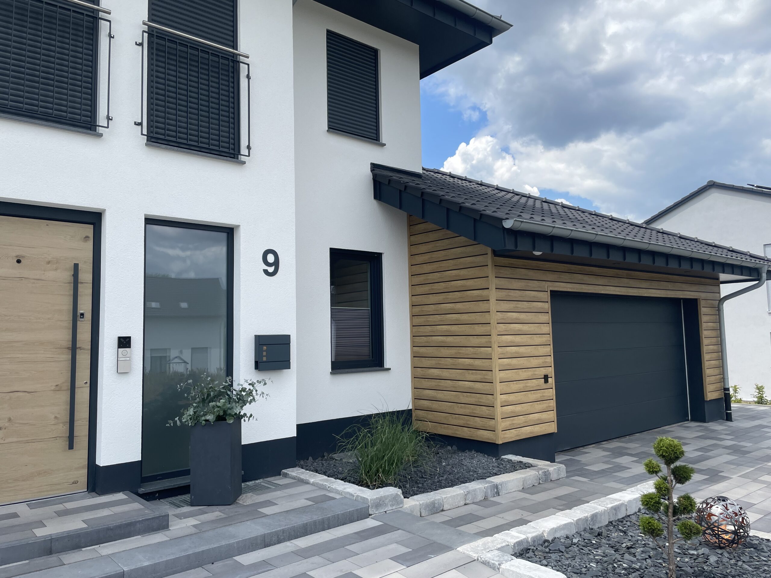 Moderne Doppelhaushälfte mit weißem Putz und anthrazitfarbenen Fenstern, einer Holzbalkonverkleidung, einer anthrazitfarbenen Hausnummer 9, einer hölzernen Eingangstür, einer großen Garage mit Holzverkleidung und einem gepflegten Vorgarten mit geometrischen Pflastersteinen und Pflanzen.