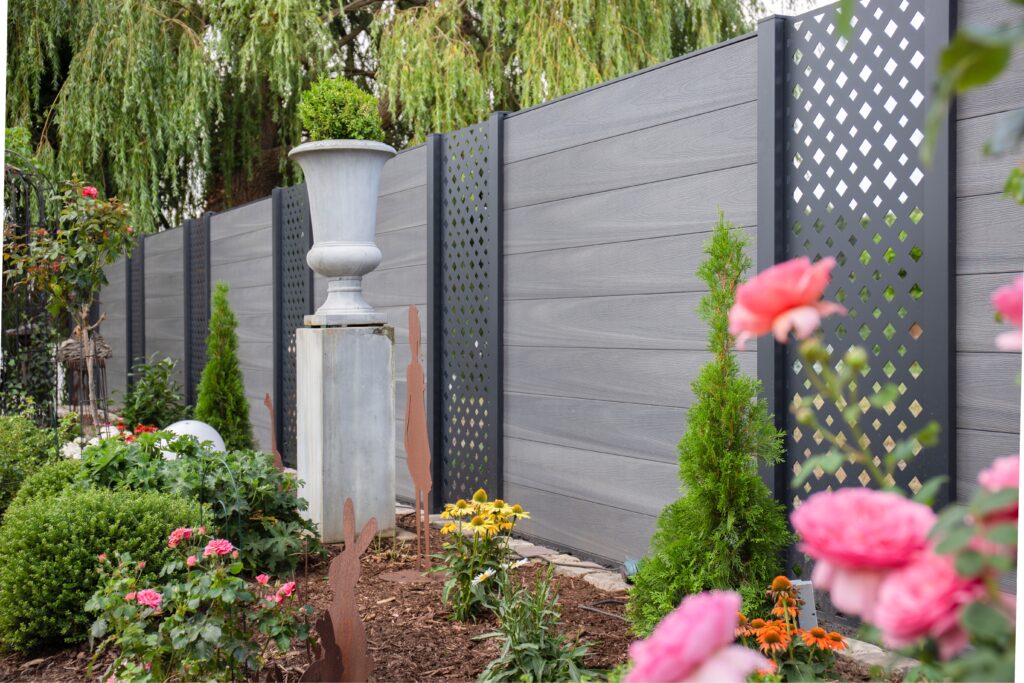 Garten mit grauem Sichtschutzzaun, verziert mit rankenden Rosen, kleinen Nadelbäumen und einer Steinvase auf einem Sockel, umgeben von grüner Landschaft.