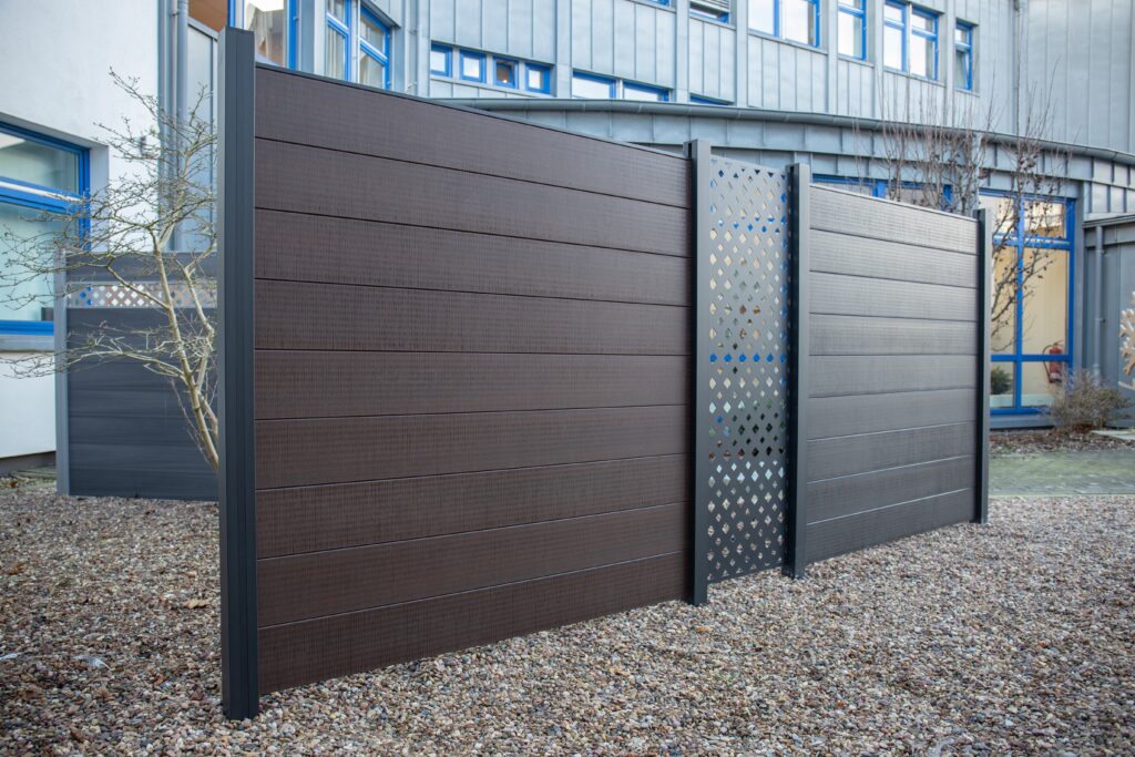 Ein moderner Zaun mit dunklem Holzdesign und einem metallischen Gitterelement in der Mitte, auf einer Kiesoberfläche vor einem Gebäude mit blauen Fensterrahmen.