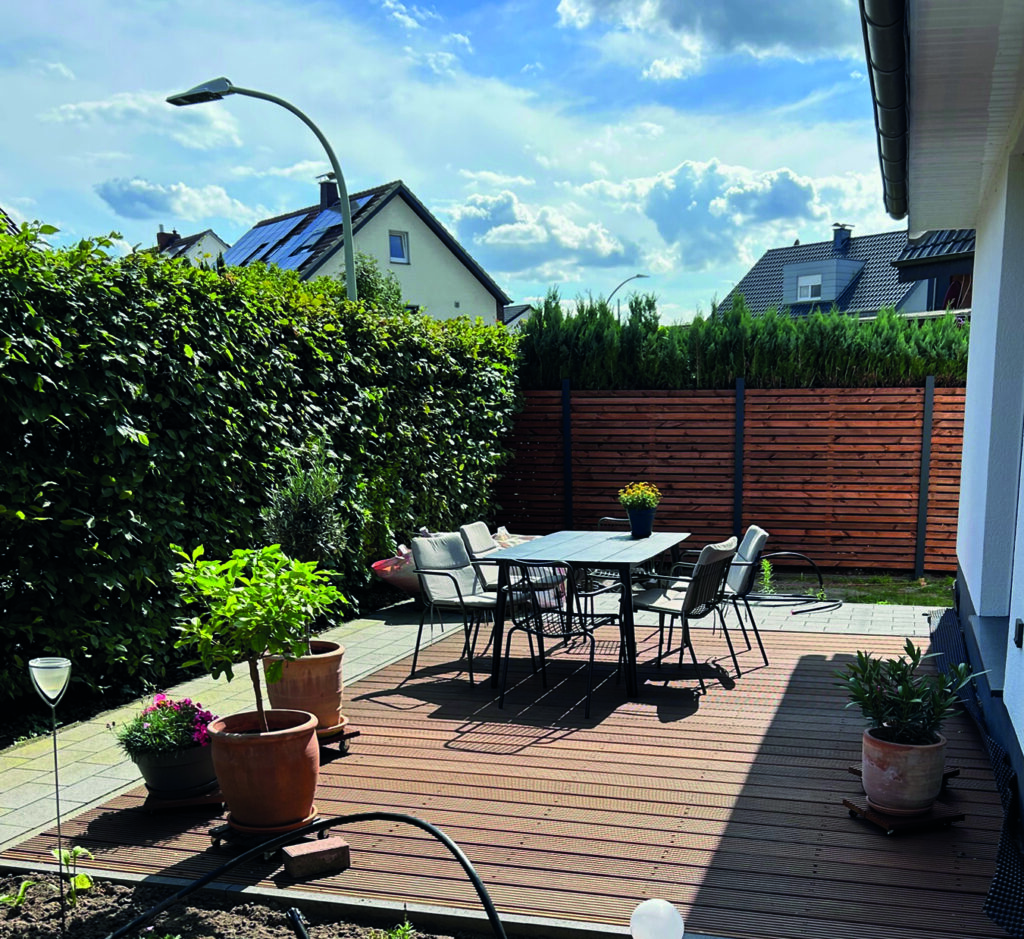 Eine Terrasse mit Holzboden und einem modernen Tisch mit Stühlen, umgeben von Pflanzkübeln und Holzzaun; im Hintergrund sind ein dicht bewachsener Garten und ein Haus zu sehen.