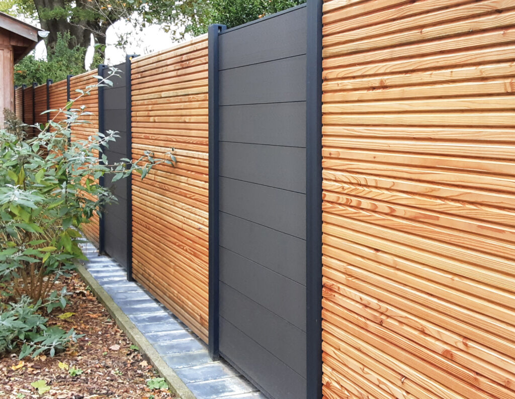 Ein Zaun mit abwechselnden Elementen aus horizontal angeordneten Holzlatten und glatten, dunkelgrauen Paneelen, daneben eine Pflanze auf einer mit Blättern bedeckten Fläche.
