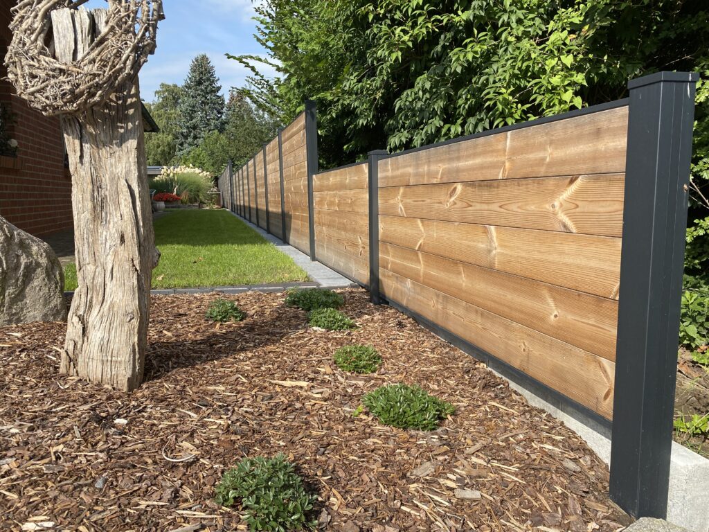 Holzzaun mit schwarzen Pfeilern verläuft entlang eines gepflegten Gartens; im Vordergrund liegt ein mit Mulch bedecktes Beet mit kleinen grünen Pflanzen, daneben ein dekorativer trockener Holzstamm.