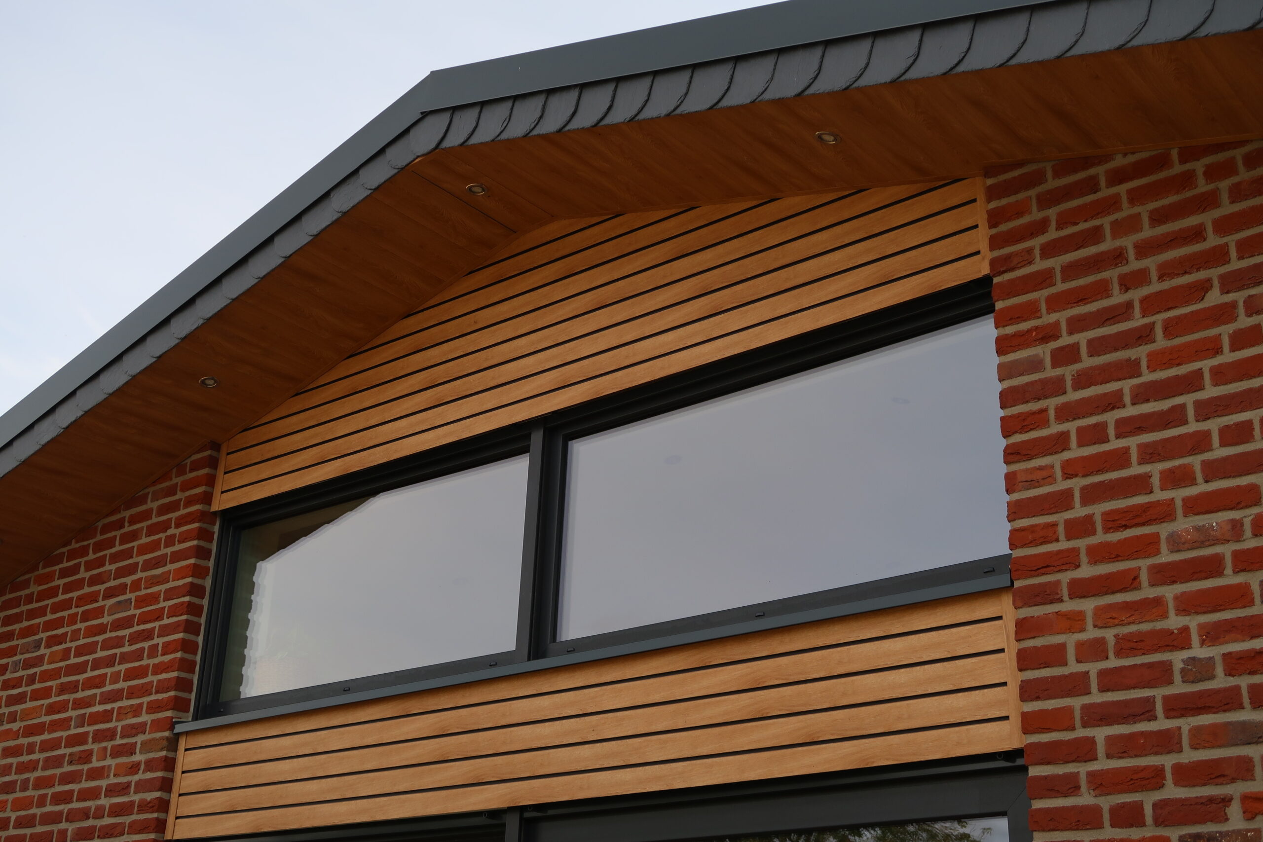 Detail eines Daches mit Holzverkleidung und sichtbaren Fenstern, daneben eine rote Ziegelwand.