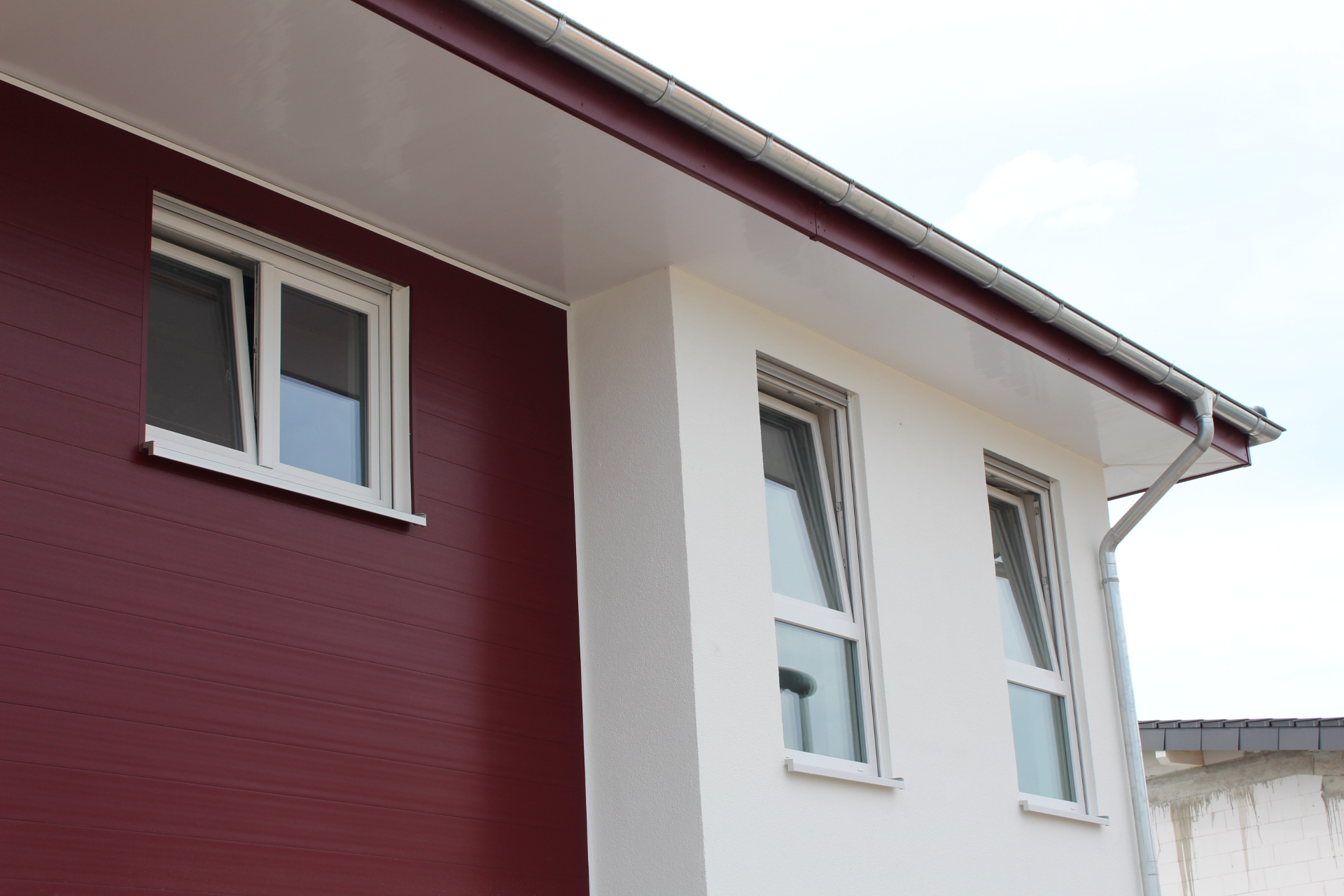 Zwei Fenster in einer zweifarbigen Gebäudefassade; links rotes Paneel, rechts weiße Putzoberfläche, mit einem grauen Dach und Regenrinne.