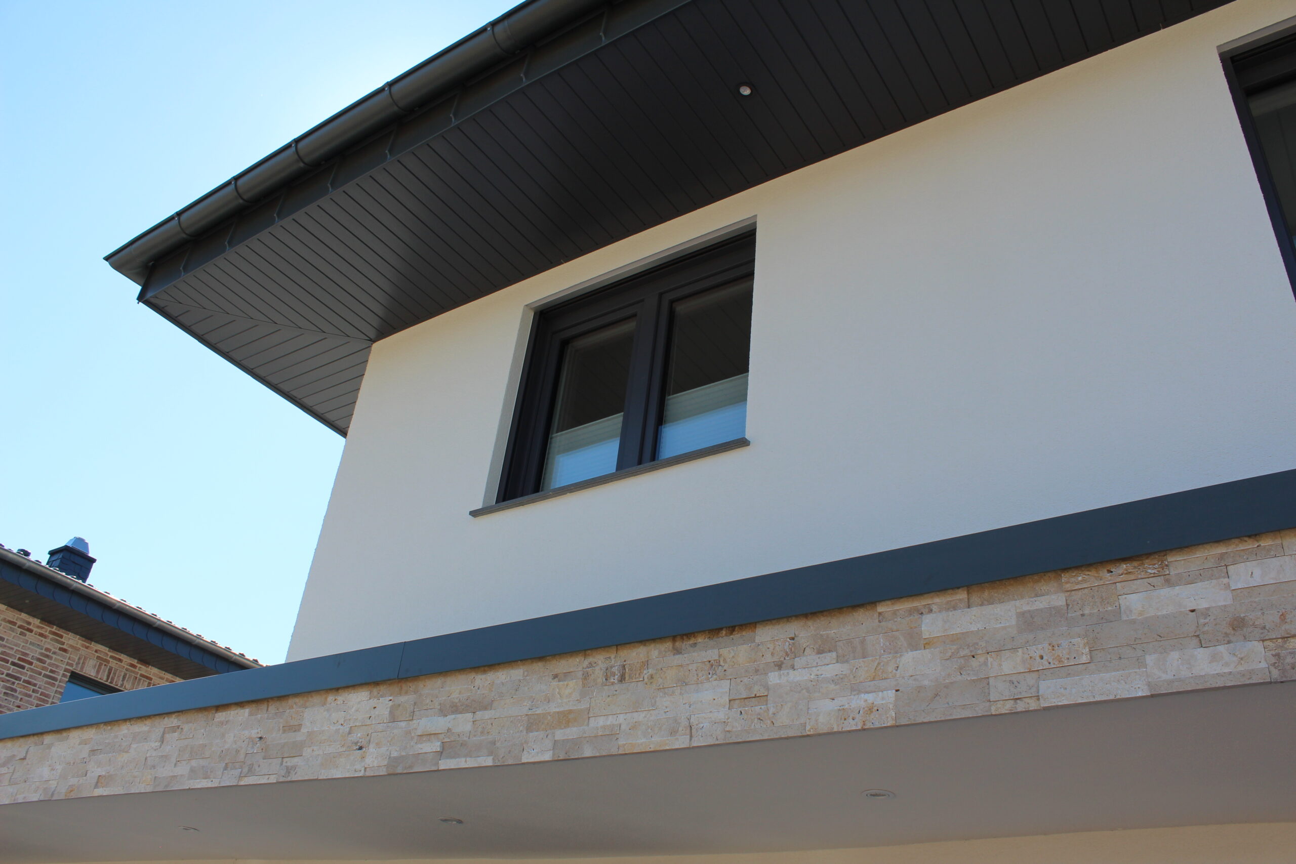 Moderne Hausfassade mit weißem Putz, grauem Dachrand und Steinverkleidung, zeigt ein Fenster im oberen Stockwerk.
